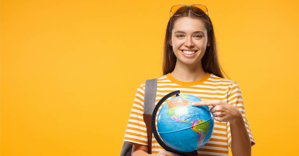 A girl wearing a striped orange shirt and standing in front of an orange background holding a globe and pointing at the U.S.