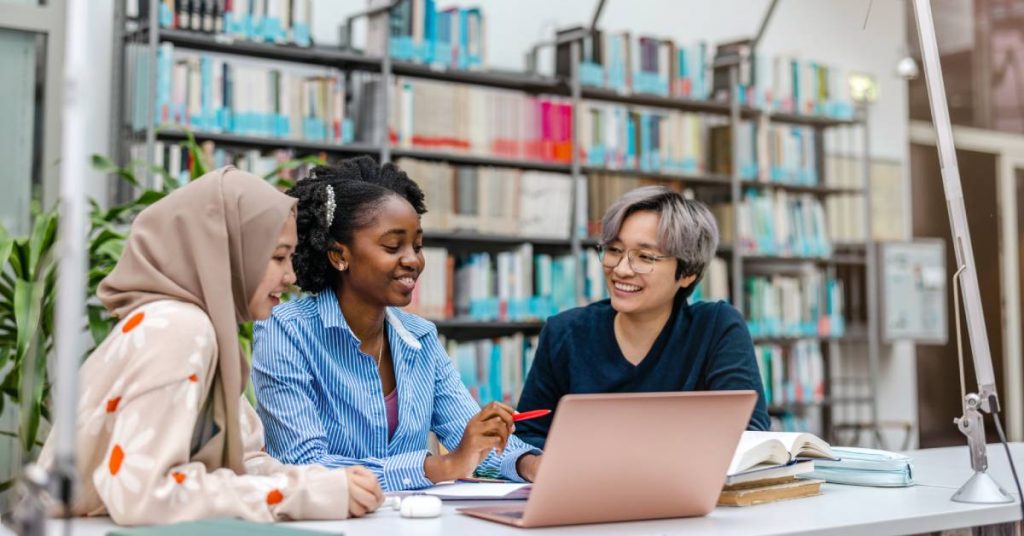 A multiethnic group of students sitting at a library table with books in the background. They are engaging in conversation.