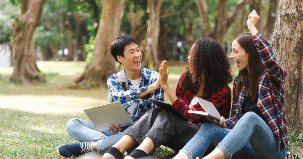 Three students sit on some grass next to a tree. Two of them are high-fiving each other while the other pumps her fist in the air.