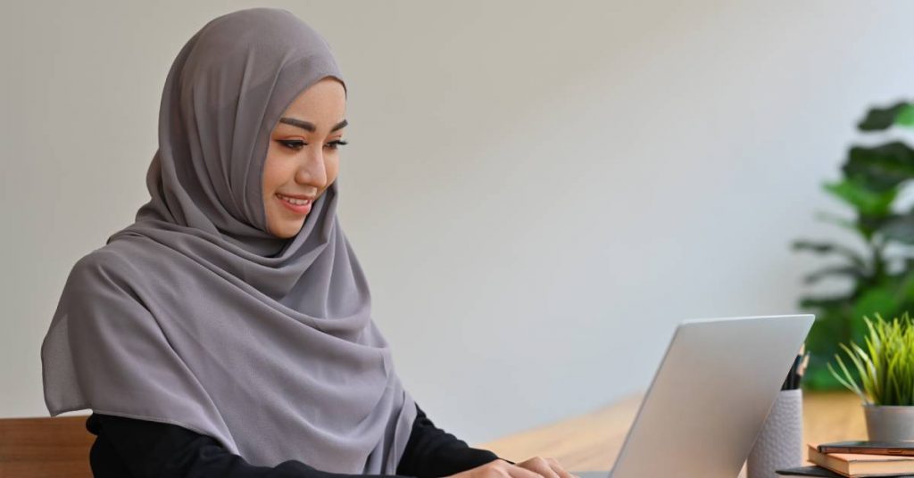 A young woman wearing a grey hijab is smiling and typing on her laptop. A pen and some notebooks are next to her.