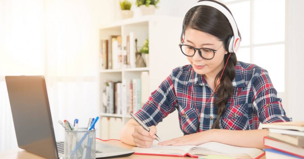 A student sitting at a desk with an open laptop and wearing headphones. She is reading a book with a pen in hand.