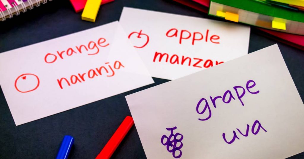 Three vocabulary cards with books in the background. The cards say "grape," "orange," and "apple" in English and Spanish.