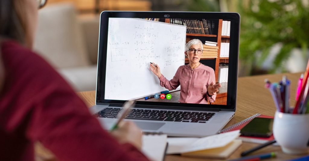 The backside of a person sitting at a table with an open laptop. The screen displays a teacher pointing at a whiteboard.