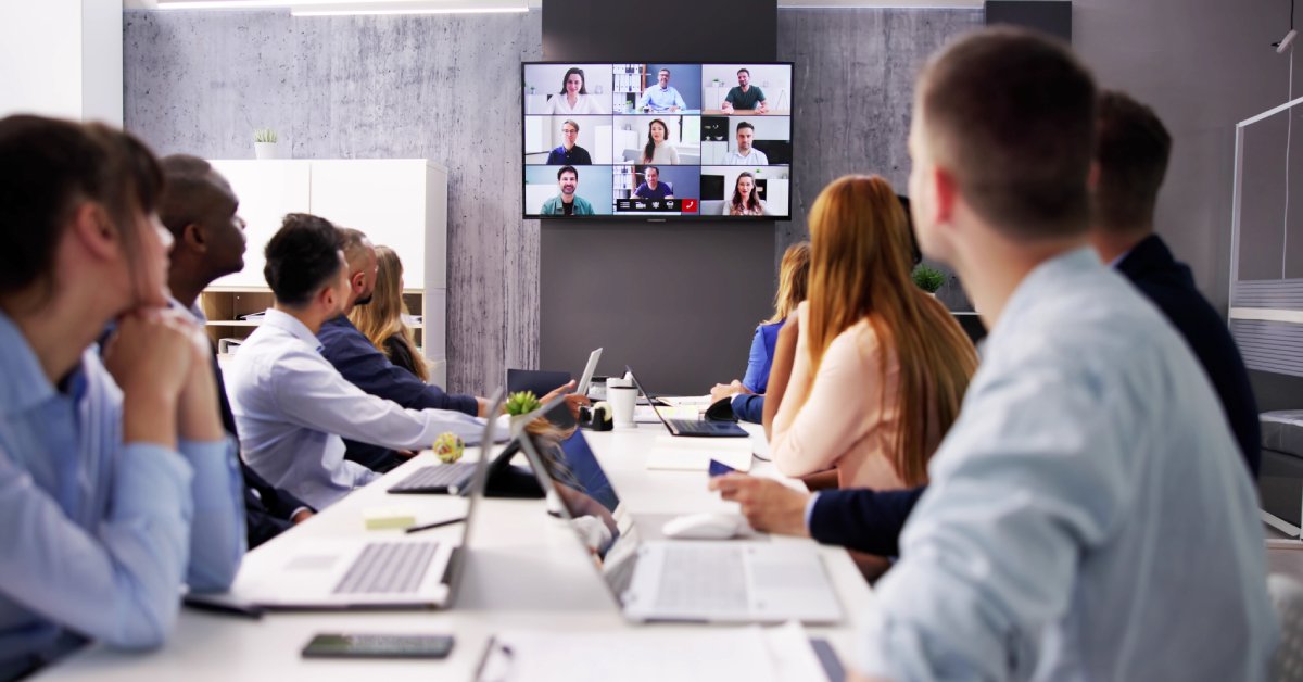 A group of people sit at a table with laptops open. They watch a screen on the wall displaying a video call.