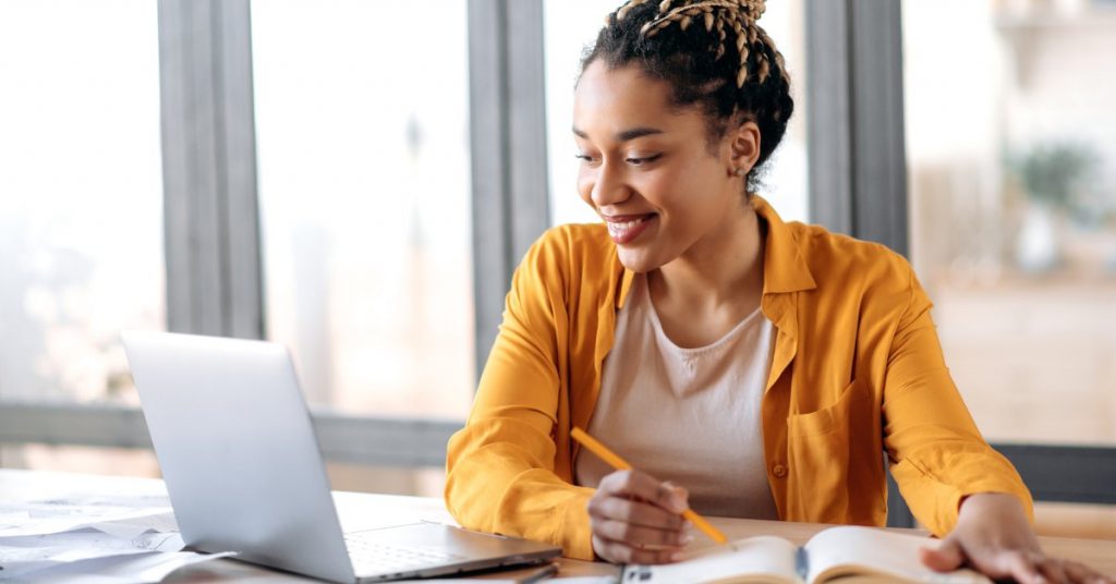 An African-American woman sits at a table with a laptop and notebooks in front of her. She holds a pencil and smiles.