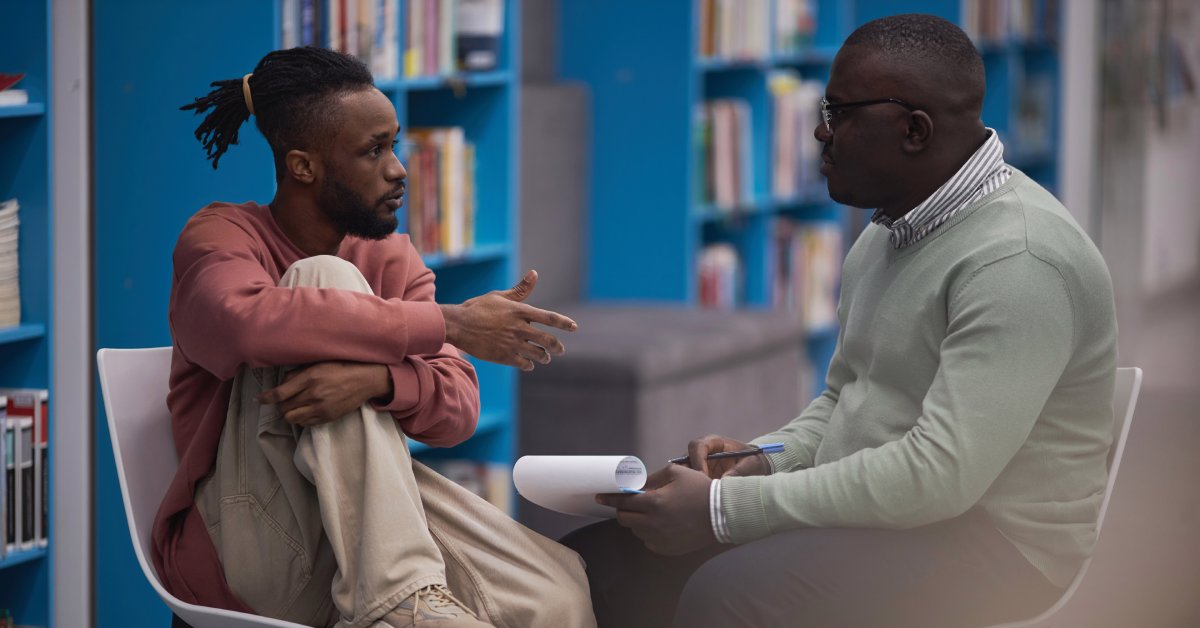 A side view of a student and an older adult speaking to each other in a library. The older man holds a clipboard and pen.