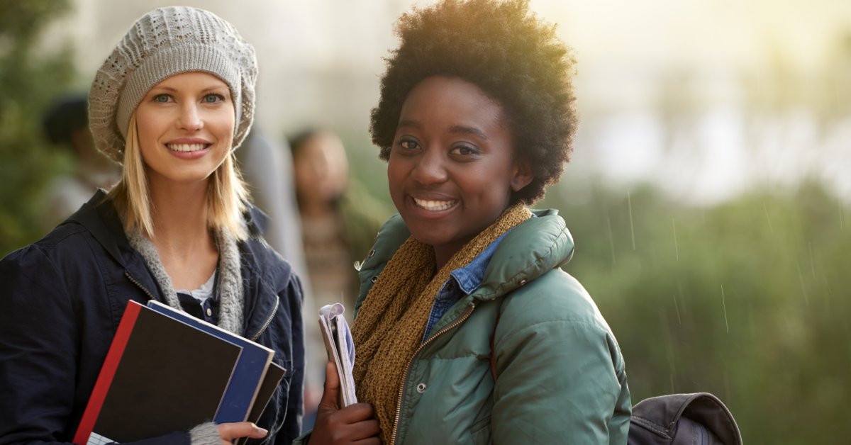 Two students wearing winter clothing smile. Both women are holding books, and one has a backpack on.