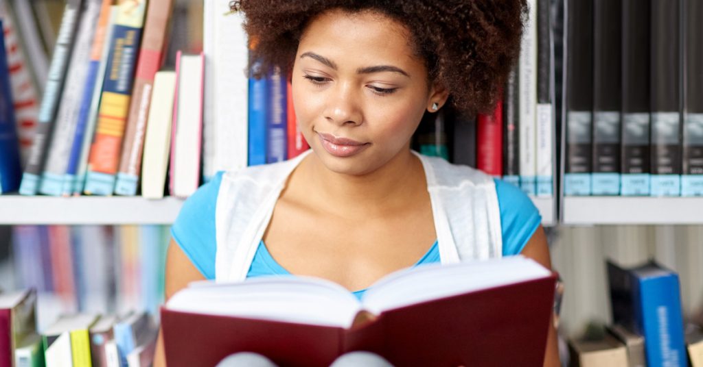 A girl sitting in a library with her back to a bookshelf. She is reading a book that is open in her lap.