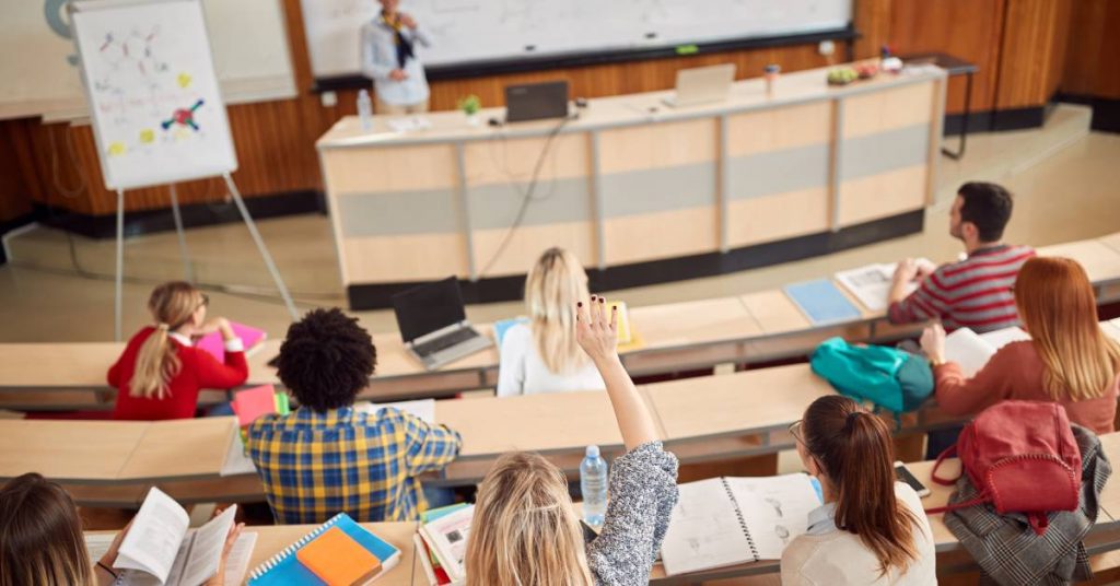 Multiple students sit in rows inside a lecture hall. There is a professor at the front of a classroom with a whiteboard behind them.