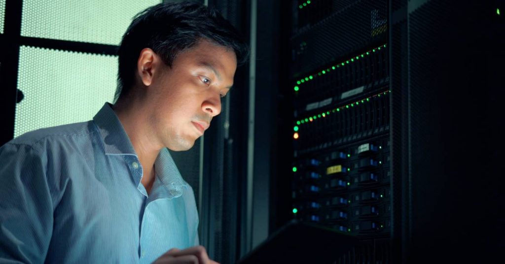 A man is in a dark room with his face illuminated by a tablet. A frosted window and a computer server are behind him.