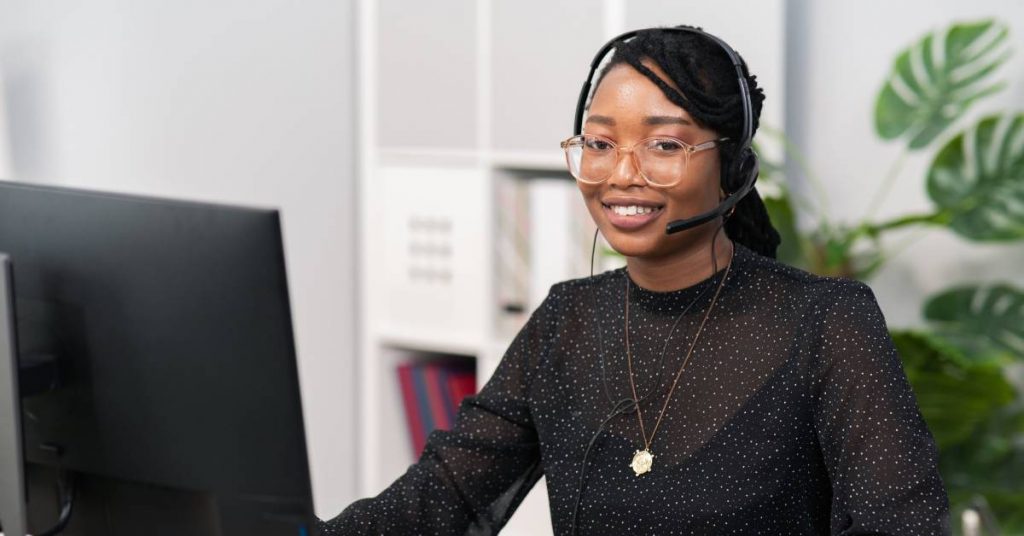 A woman sits at a desk with a bookshelf and plant behind her. She is wearing headphones and smiling.