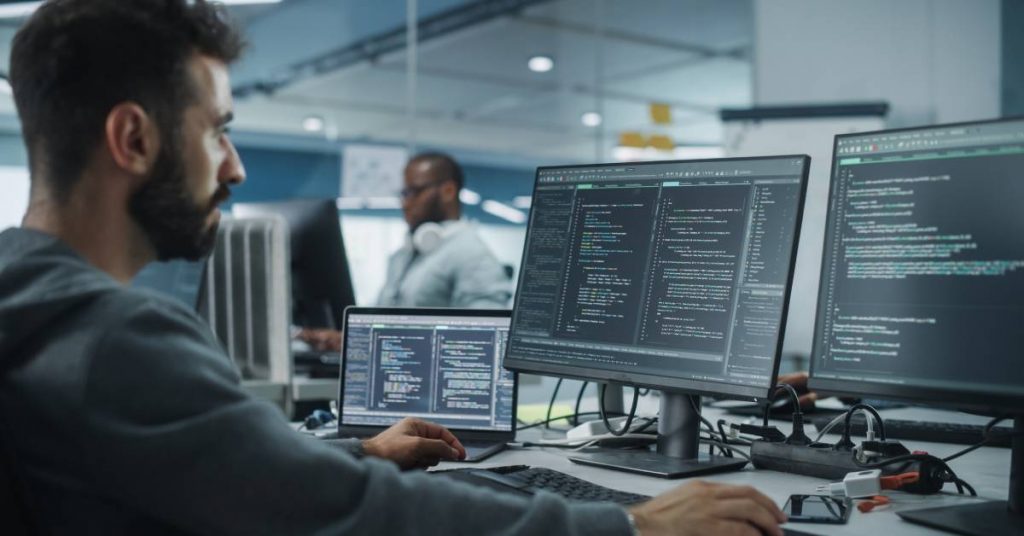 A man sits at a computer desk in an office. There are three different screens in front of him with coding on them.