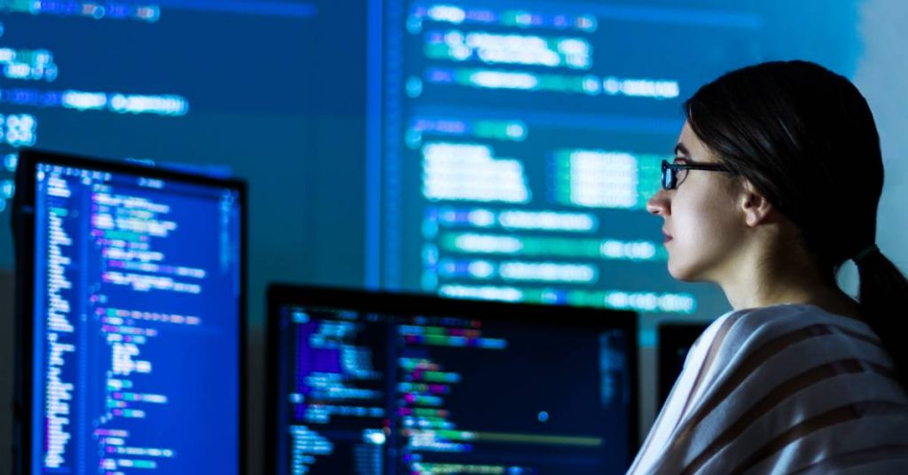 A woman wearing glasses and a striped shirt is sitting in front of large computer screens. She is looking at different coding.