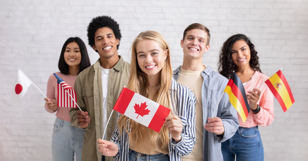 Individuals of different ethnicities stand together as they smile and hold flags of several countries.