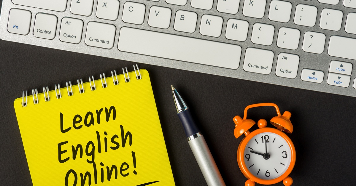 A yellow notepad stating "learn English online," next to a pen and a small clock underneath a keyboard.