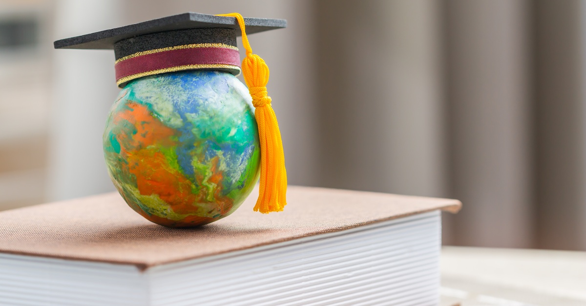 A black and red master degree cap and gold tassel adorn a globe as the globe sits on a thick book on a surface.