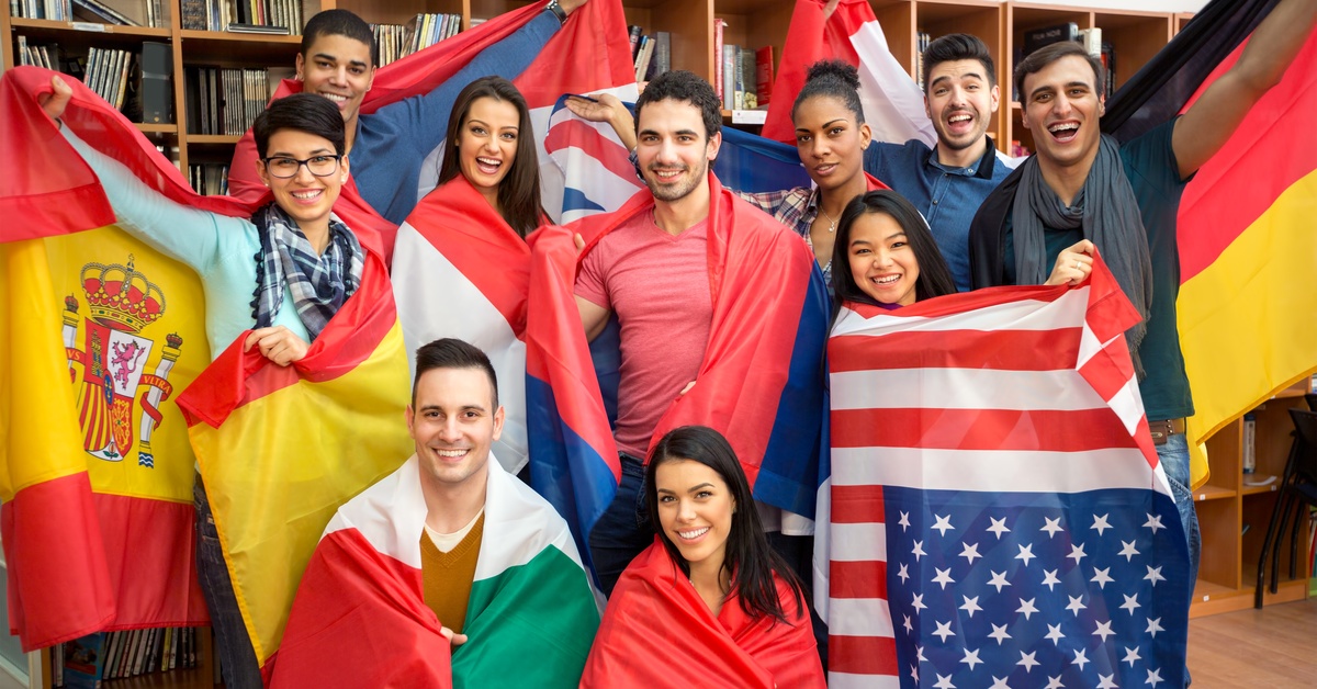 Individuals of different ethnicities, with various countries' flags wrapped around them, stand in front of bookcases.