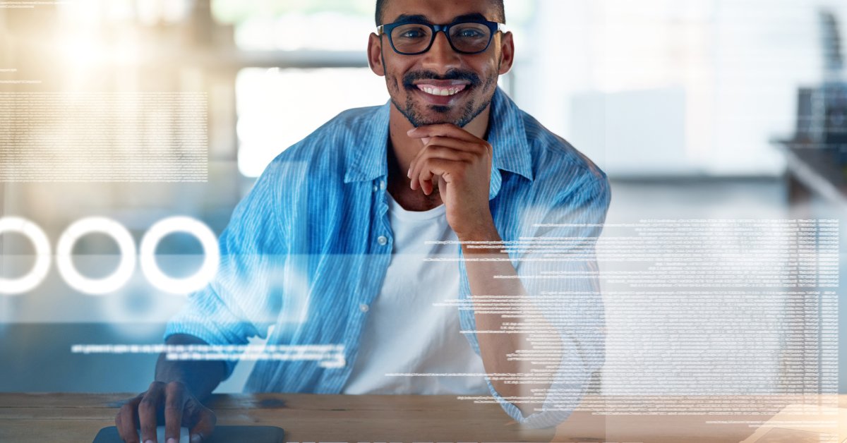 A man smiling as he looks ahead. His left hand is against his chin and his right hand is on a computer mouse.